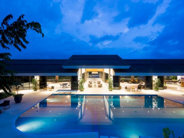 A luxurious modern building with a large illuminated swimming pool in the foreground, surrounded by lounges and trees under a blue sky.