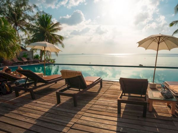 A serene poolside scene with lounge chairs, umbrellas, and a view of the ocean, bordered by palm trees under a partly cloudy sky.