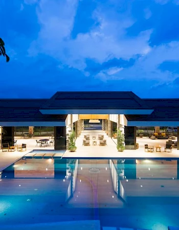 A modern building with a lit interior, featuring an outdoor pool reflecting the structure against a twilight sky.