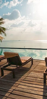 A tranquil poolside scene with lounge chairs, umbrellas, and palm trees overlooking a serene body of water under a partly cloudy sky.