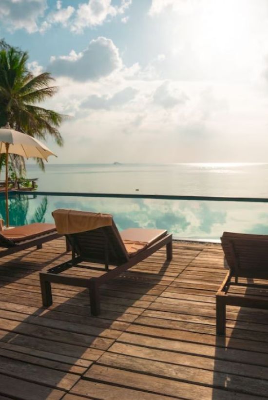 A serene poolside view with sun loungers, umbrellas, and a tranquil ocean in the background under a partly cloudy sky.