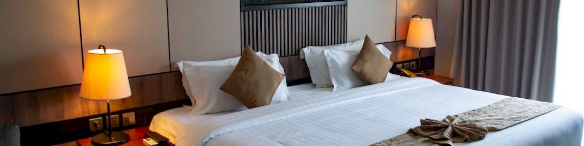 A cozy hotel room featuring a king-sized bed with pillows, warm lighting, wall art, and a neat decor, creating a relaxing atmosphere.
