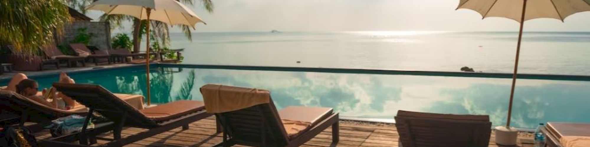 A serene poolside setup with lounge chairs, umbrellas, and a scenic ocean view at sunset, surrounded by palm trees.