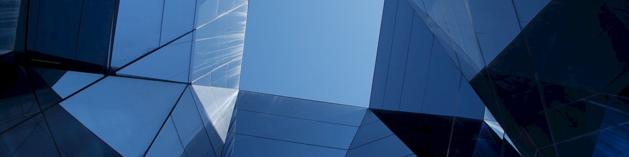 The image shows tall, reflective glass buildings forming an angular, open atrium with a clear blue sky visible above.