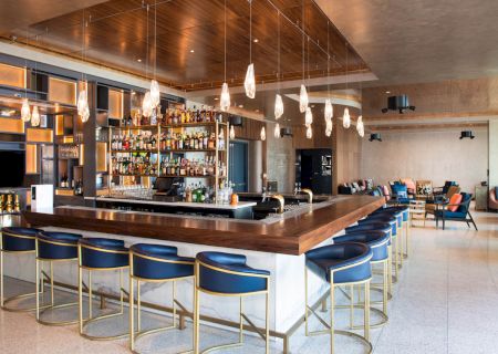 A modern bar with blue stools, a wooden countertop, and pendant lights. It features a fully stocked back wall and comfortable seating area.