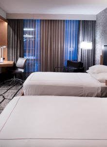 A modern hotel room with two beds, a wall-mounted TV, a desk, and a window with curtains. Stylish lighting and patterned wallpaper complete the design.