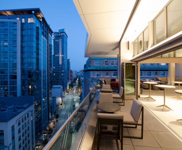 A modern rooftop terrace overlooking a city street, featuring seating areas and buildings with large windows during the evening.