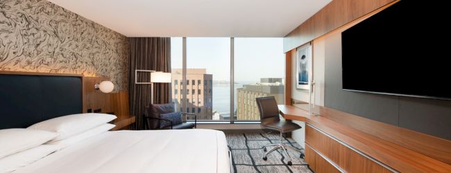 A modern hotel room with a large bed, desk, TV, and window view of buildings. The décor includes wood accents and a patterned carpet.
