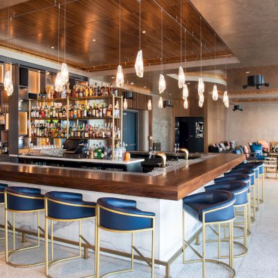 A modern bar with a wooden counter, blue stools, hanging lights, and a well-stocked shelf. Cozy seating area in the background.