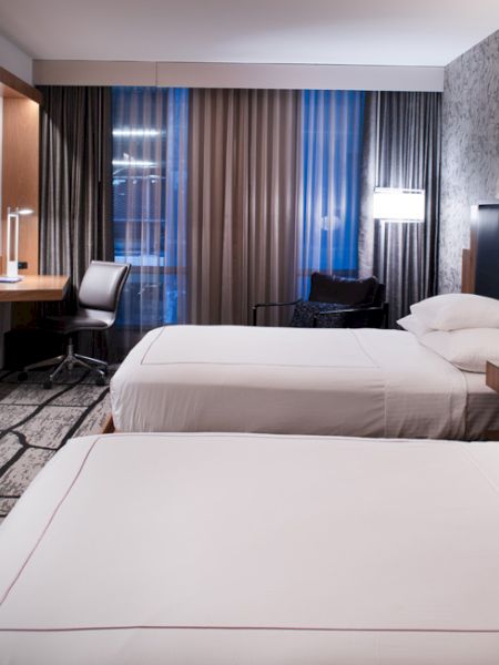 A modern hotel room with two beds, a TV, a desk, and large windows with curtains. Elegant decor includes patterned walls and floor lamps.