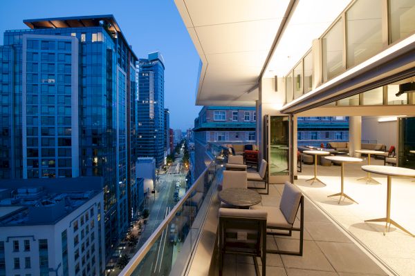 A modern balcony overlooking a cityscape at dusk, with outdoor seating and buildings lit up against a blue sky.
