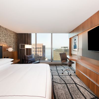 A modern hotel room with a bed, desk, chair, large TV, and window view of buildings and water, featuring stylish decor and ample lighting.