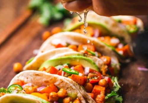 The image shows a hand squeezing lime over a row of tacos filled with chickpeas, vegetables, and garnished with cilantro on a wooden surface.