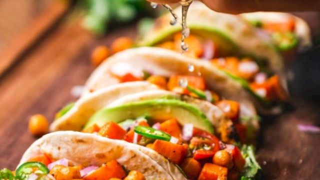 The image shows a hand squeezing lime over a row of tacos filled with chickpeas, vegetables, and garnished with cilantro on a wooden surface.