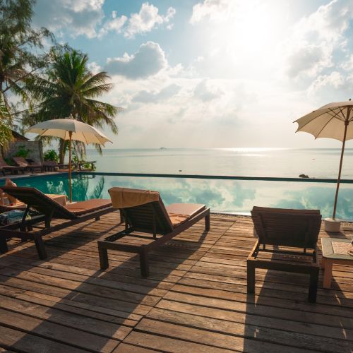 A serene tropical scene with lounge chairs, umbrellas, a pool, and palm trees by the ocean under a partly cloudy sky and bright sun.