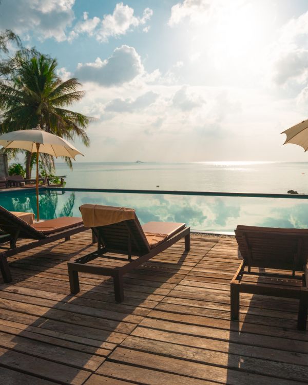 A serene tropical scene with lounge chairs, umbrellas, a pool, and palm trees by the ocean under a partly cloudy sky and bright sun.