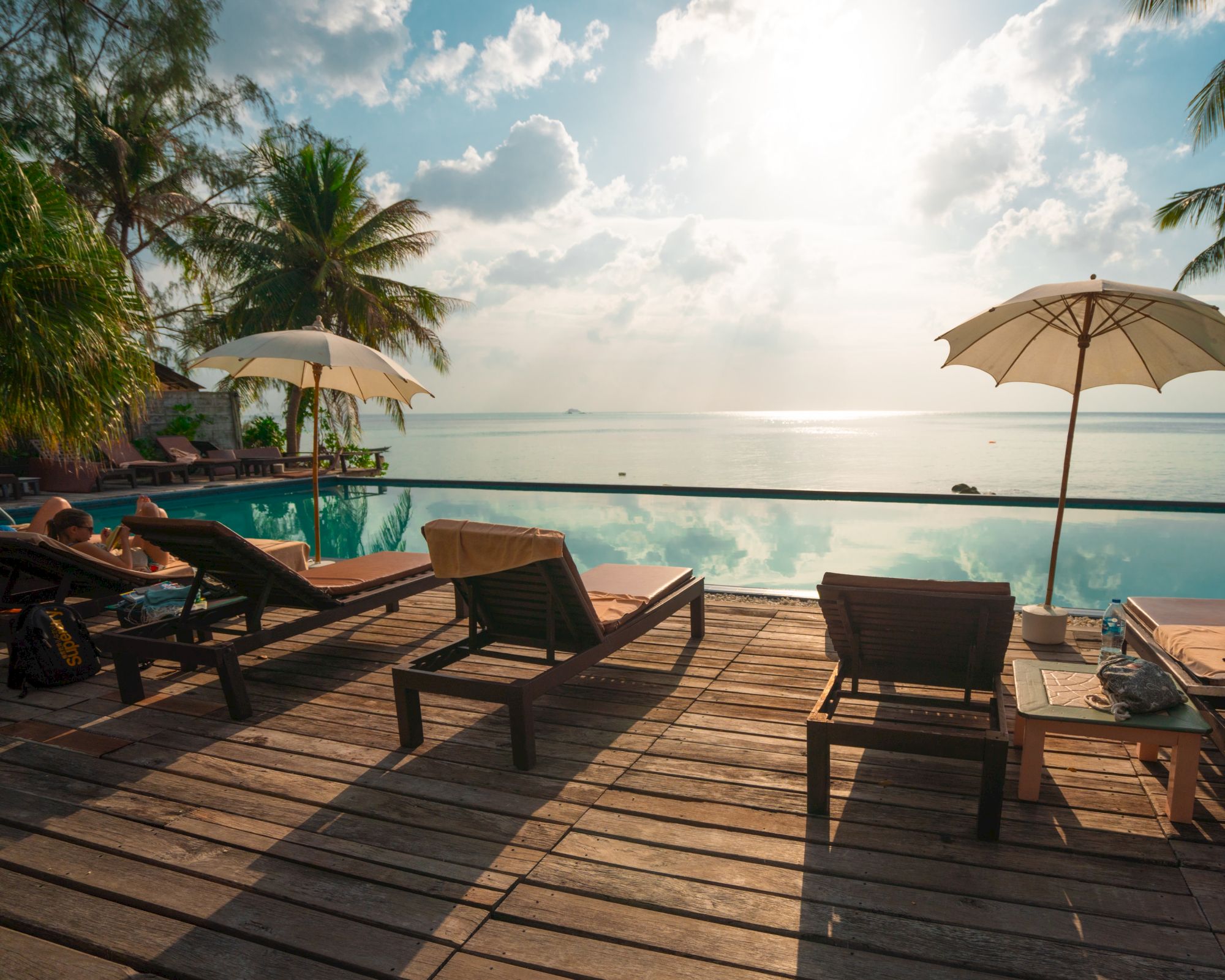 A serene beach setting with lounge chairs, sun umbrellas, and an infinity pool overlooking the ocean under a partly cloudy sky.