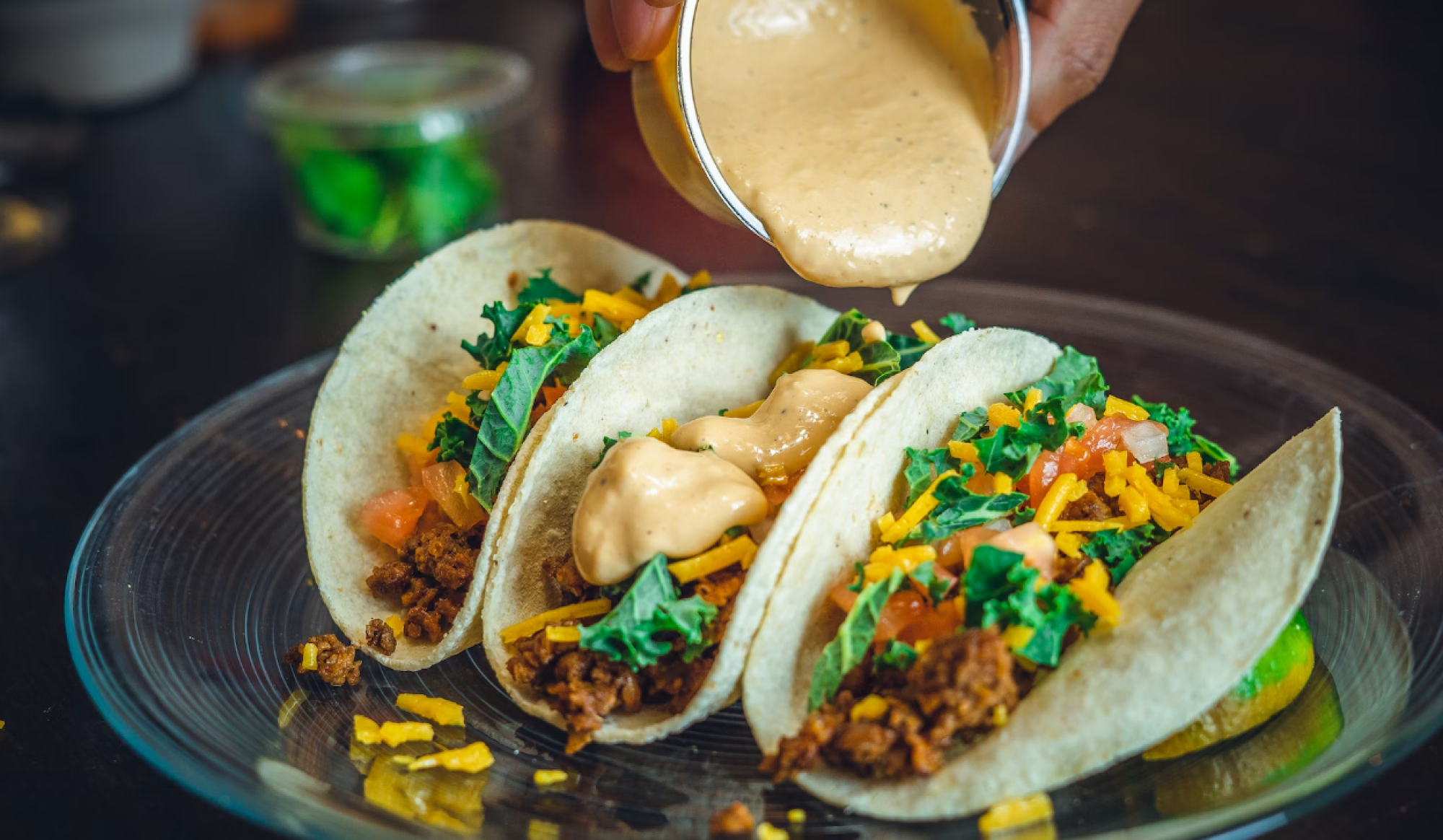 Three tacos with ground meat, cheese, tomatoes, and lettuce are on a plate, with sauce being poured over them.