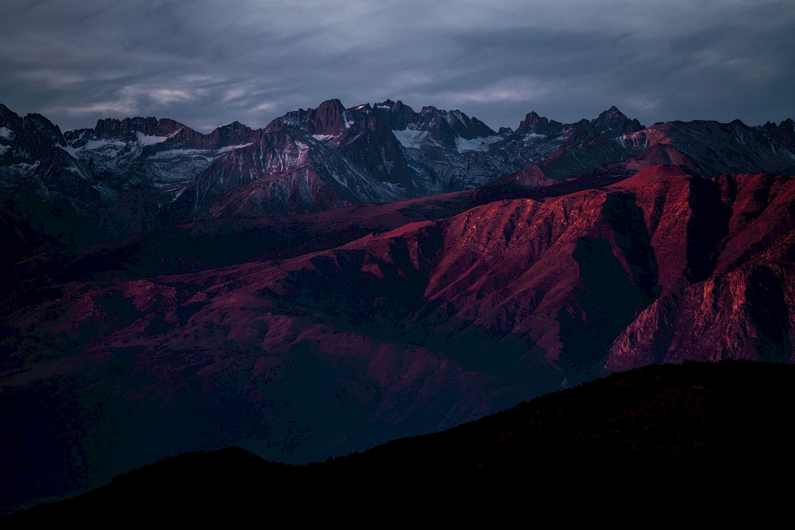 A mountain range with rocky peaks is illuminated by deep red and purple hues during sunset, under a cloudy sky.