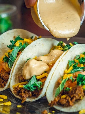 Three tacos with ground meat, cheese, lettuce, and sauce being poured on top.