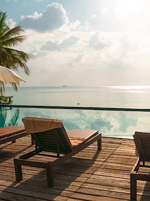 Lounge chairs and umbrellas by a pool overlook a serene ocean view, with palm trees and a sunlit sky creating a relaxing atmosphere.
