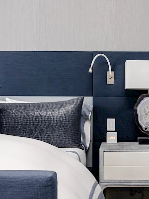 The image shows a modern bedroom with a navy blue headboard, white bedding, a lamp on a nightstand, and grey and blue decorative pillows.