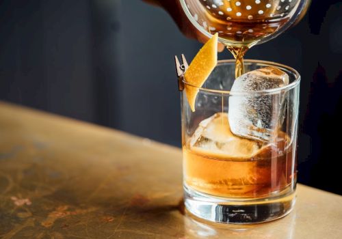 A hand pours a brown drink into a glass with ice and a lemon twist garnish on a countertop.