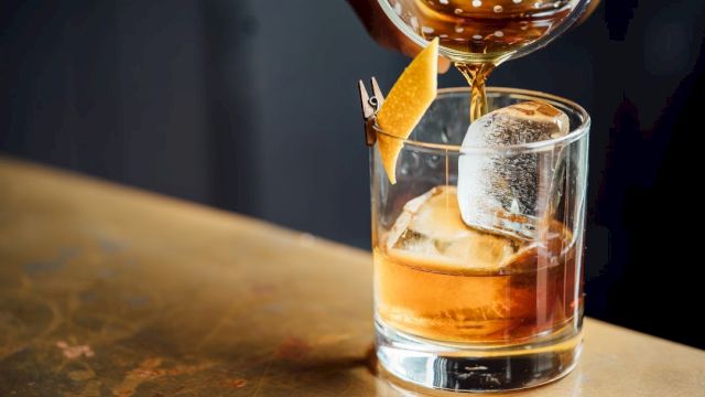 A hand pours a brown drink into a glass with ice and a lemon twist garnish on a countertop.