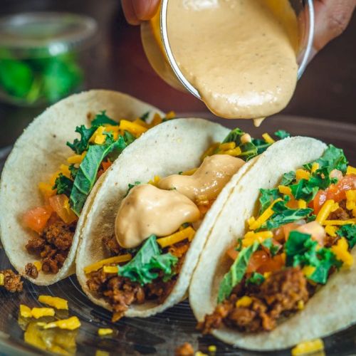 Three tacos filled with ground meat, shredded cheese, tomatoes, and greens, with sauce being poured over them.