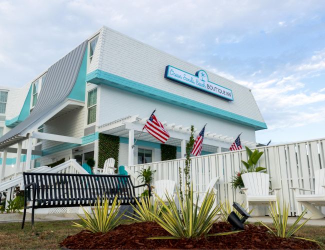 The image shows a two-story building with a sign, featuring American flags, outdoor seating, and landscaping in front.