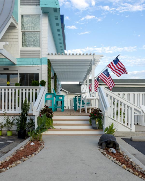 A house entrance with a white pergola, teal chairs, and multiple flags beside the pathway, surrounded by plants and blue sky above.