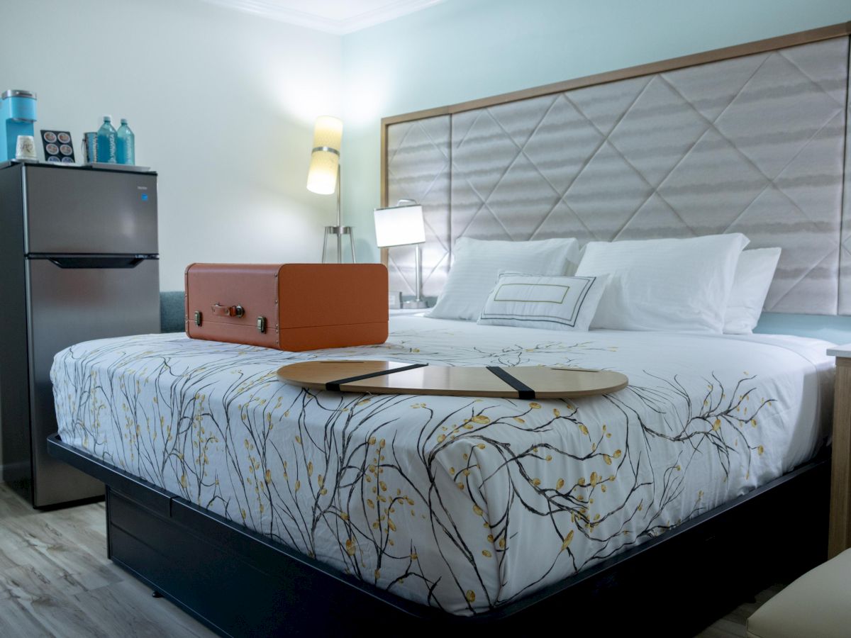 A cozy bedroom with a patterned bedspread, an orange suitcase, a tray on the bed, a mini fridge, and soft lighting.