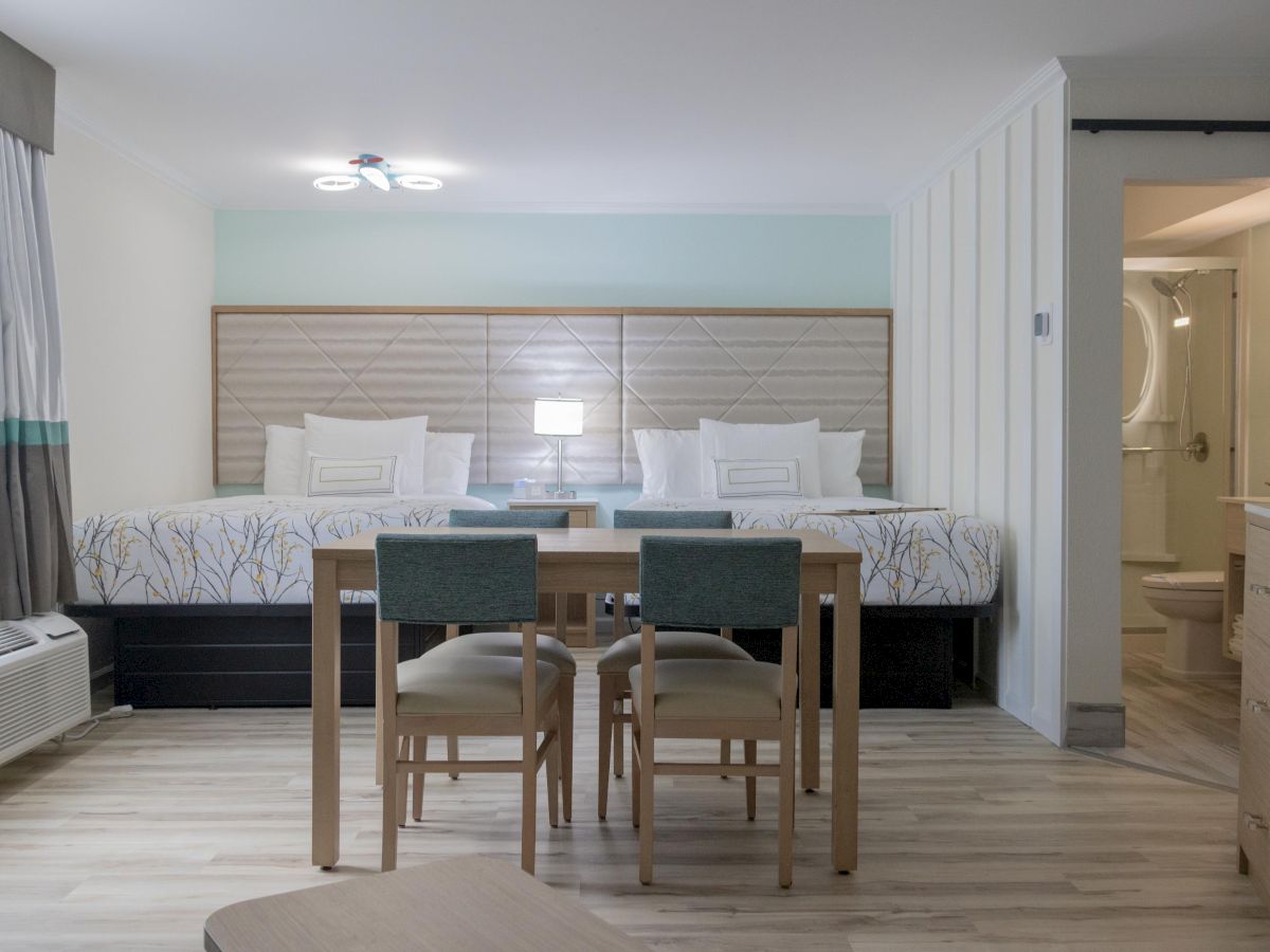 The image shows a modern hotel room with two beds, a table with chairs, and a bathroom in the corner.