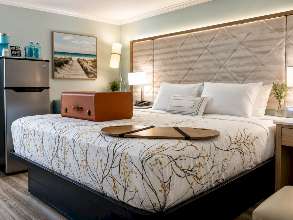 A modern hotel room with a made bed, pillows, lamps, suitcase, fridge, and drinks.