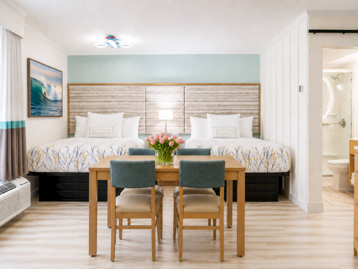 Bright hotel bedroom with two beds, floral artwork, a small table with chairs, and an en-suite bathroom visible through the doorway.