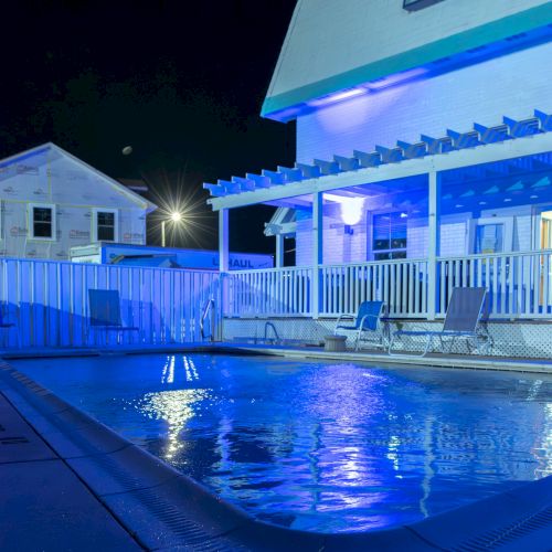 A night view of a swimming pool with blue lighting, surrounded by a house with a pergola and outdoor seating.