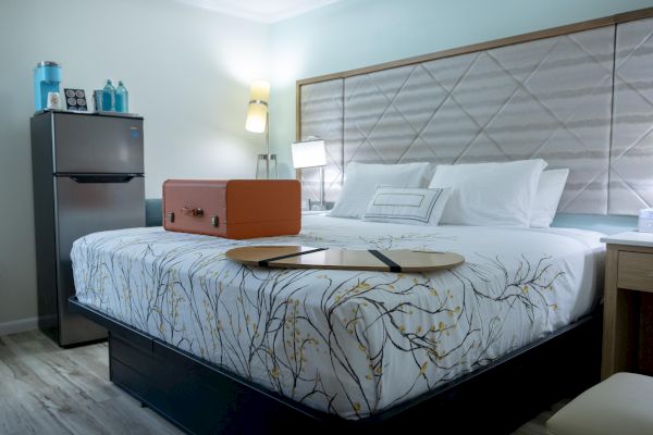 A cozy hotel room with a neatly made bed, an orange suitcase, a small fridge, and decorative items on a nightstand.
