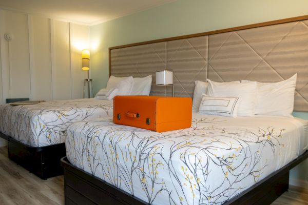 The image shows a hotel room with two beds, floral bedding, a bright orange suitcase on a bed, and modern lighting.