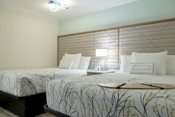 The image shows a hotel room with two double beds, featuring floral-patterned bedding, a quilted headboard, and a bedside table with a lamp.