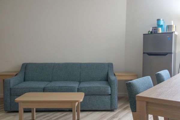 A living room with a teal sofa, wooden table, dining chairs, and a fridge topped with items in the corner.