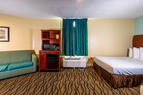 The image shows a hotel room with a bed, green sofa, wooden cabinet with a microwave, and window with blue curtains.