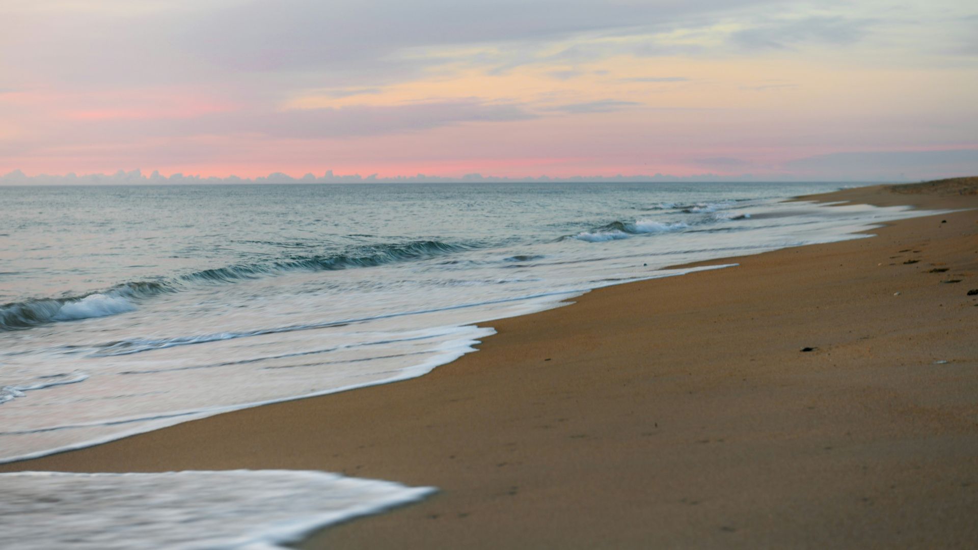 A serene beach scene with gentle waves lapping at the sandy shore under a pastel-colored sky at sunset.
