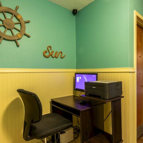 A small hallway corner with a desk, chair, computer, and printer. A ship wheel decor and "Sun" sign adorn the teal and yellow walls.