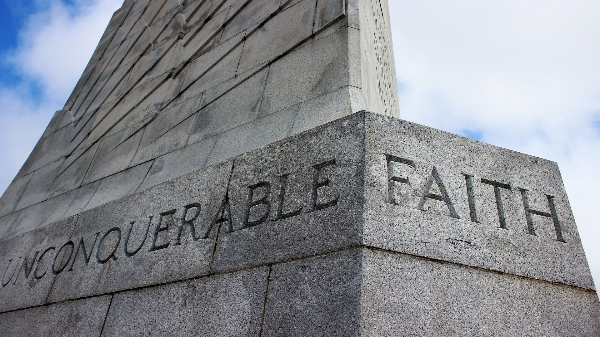 A stone monument with the words "UNCONQUERABLE FAITH" engraved on it, with a partly cloudy sky in the background.