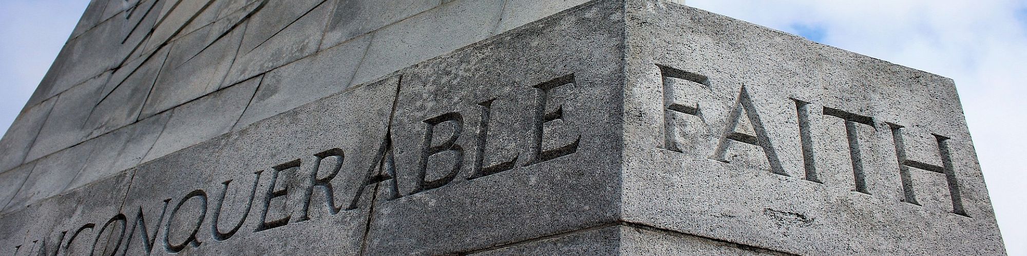 A stone monument with the words "UNCONQUERABLE FAITH" engraved on it, with a partly cloudy sky in the background.