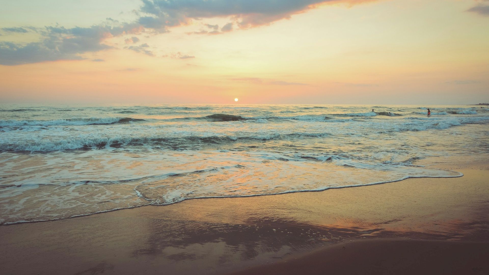 A serene beach scene at sunset with calm waves gently hitting the shore, under a sky painted in soft orange and pink hues.