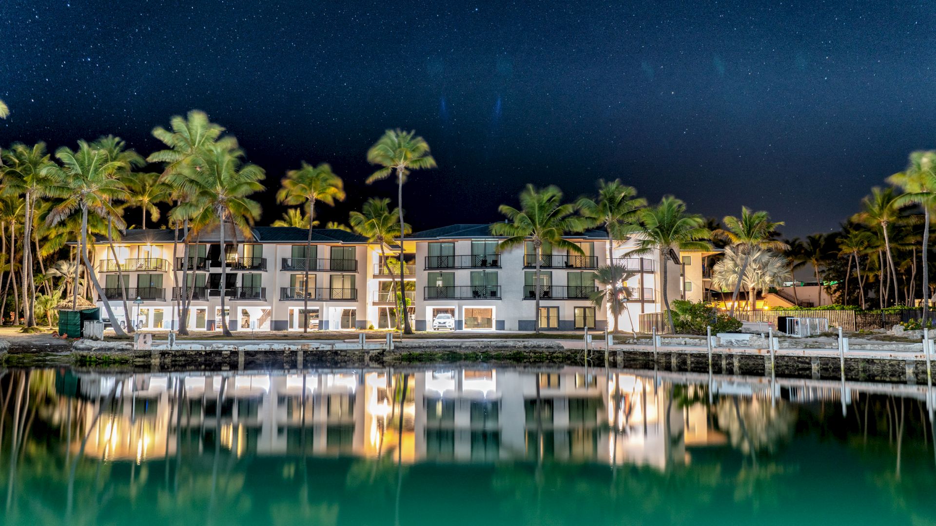 A luxurious waterfront hotel under a starry night sky, surrounded by palm trees, perfectly reflected in the calm water, creating a serene scene.