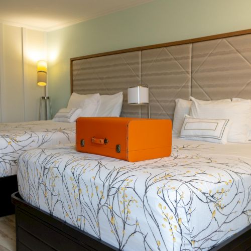 The image shows a hotel room with two beds, an orange suitcase on one, and elegant bedding with a nature-inspired pattern.