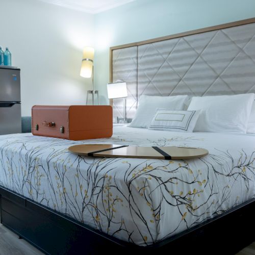A neatly arranged bedroom with a double bed, fridge, and suitcase on bedspread. Lamps and decor enhance the cozy atmosphere.
