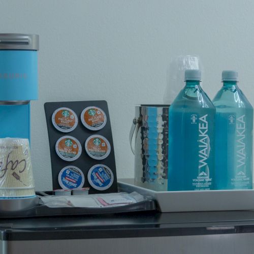 A blue coffee maker, K-cups, water bottles, and paper cups are arranged on a tabletop.